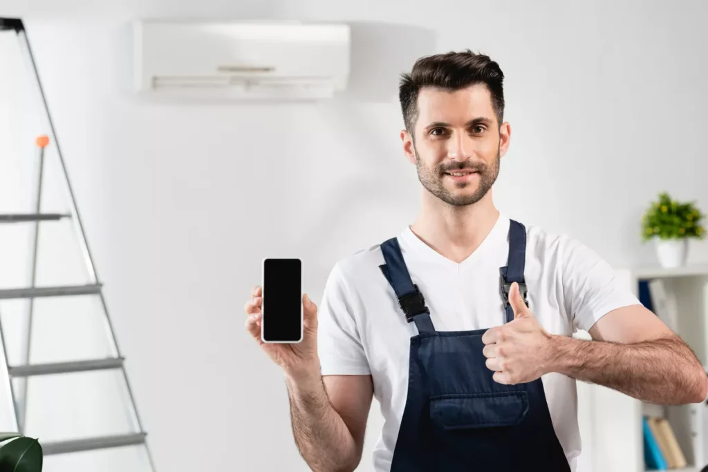 A smiling AC service technician in blue overalls giving a thumbs-up and holding a cellphone in front of a wall-mounted cooling unit.
