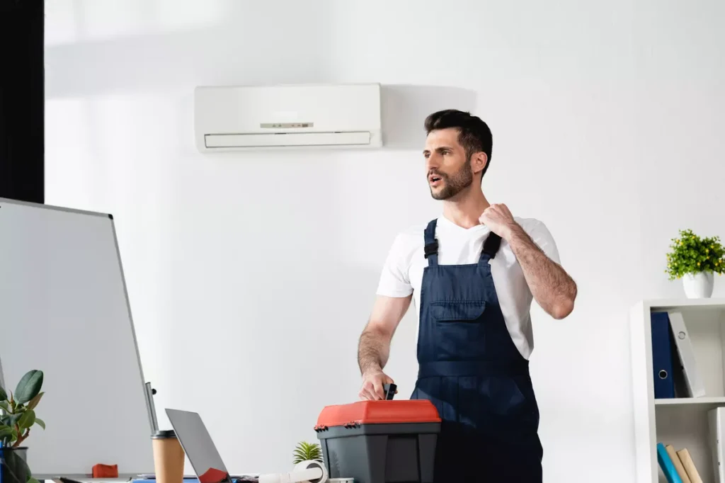 A professional HVAC technician in blue overalls standing in a bright office with a toolbox, ready to install a new air conditioning system.
