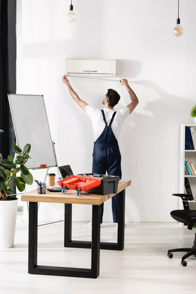An HVAC specialist in blue overalls using a measuring tape to plan an AC installation in a bright Aurora office setting.