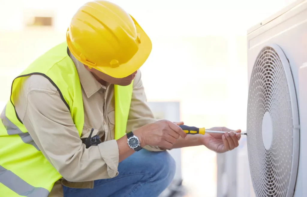 A safety-certified HVAC technician in a reflective vest using a screwdriver to repair an outdoor air conditioning condenser unit in Aurora.