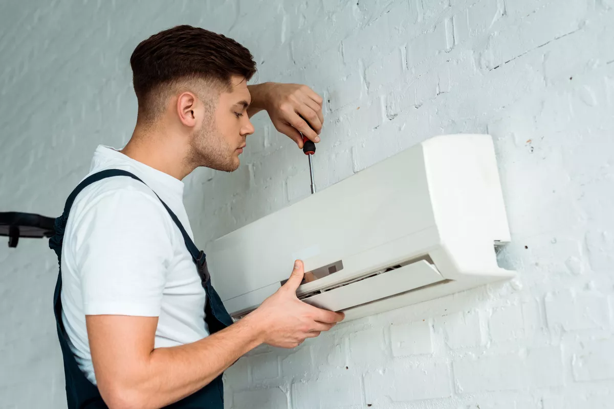 Technician in blue overalls installing a new wall-mounted air conditioning system on a white brick wall in an Aurora home.